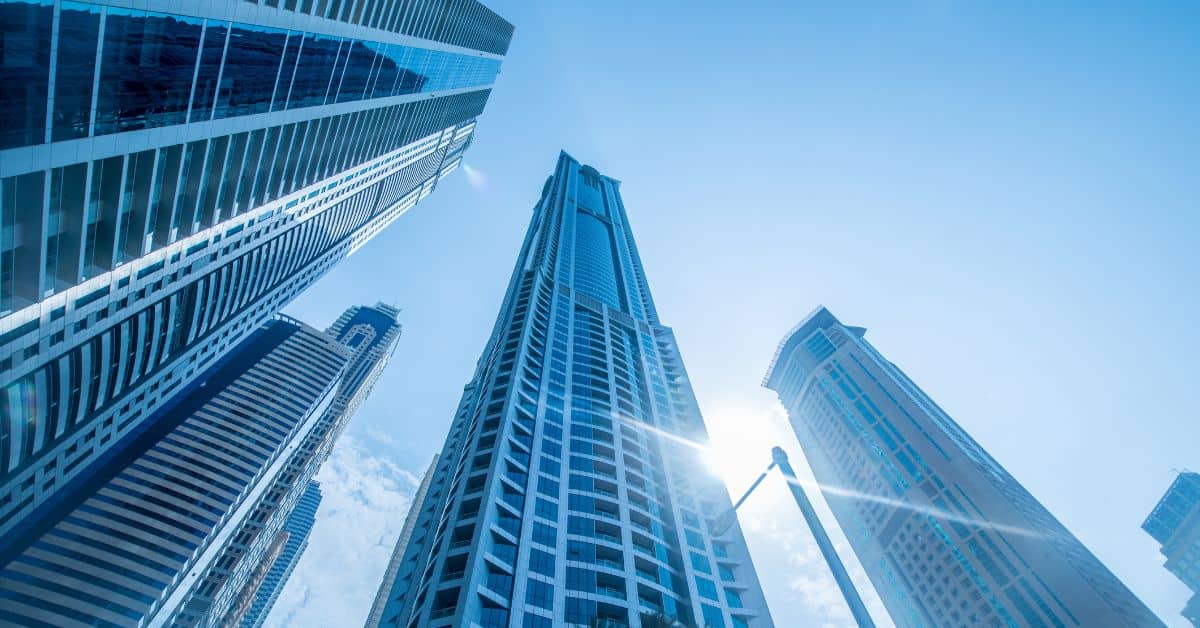 Low-angle view of multiple modern glass skyscrapers reaching into a clear blue sky, with a bright sun flare visible.