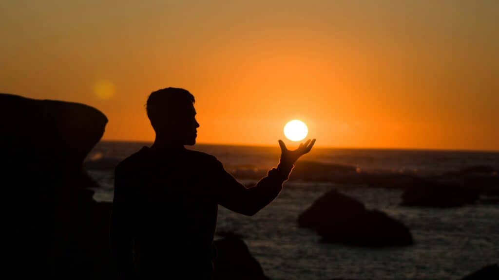 A person in silhouette appears to hold the setting sun in their hand against an orange and yellow sky over the ocean.