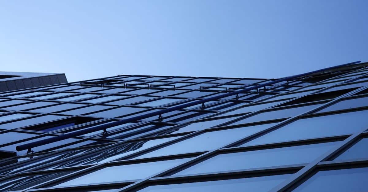 Upward view of a modern building's glass facade with a blue pipe running diagonally across it, against a clear blue sky.