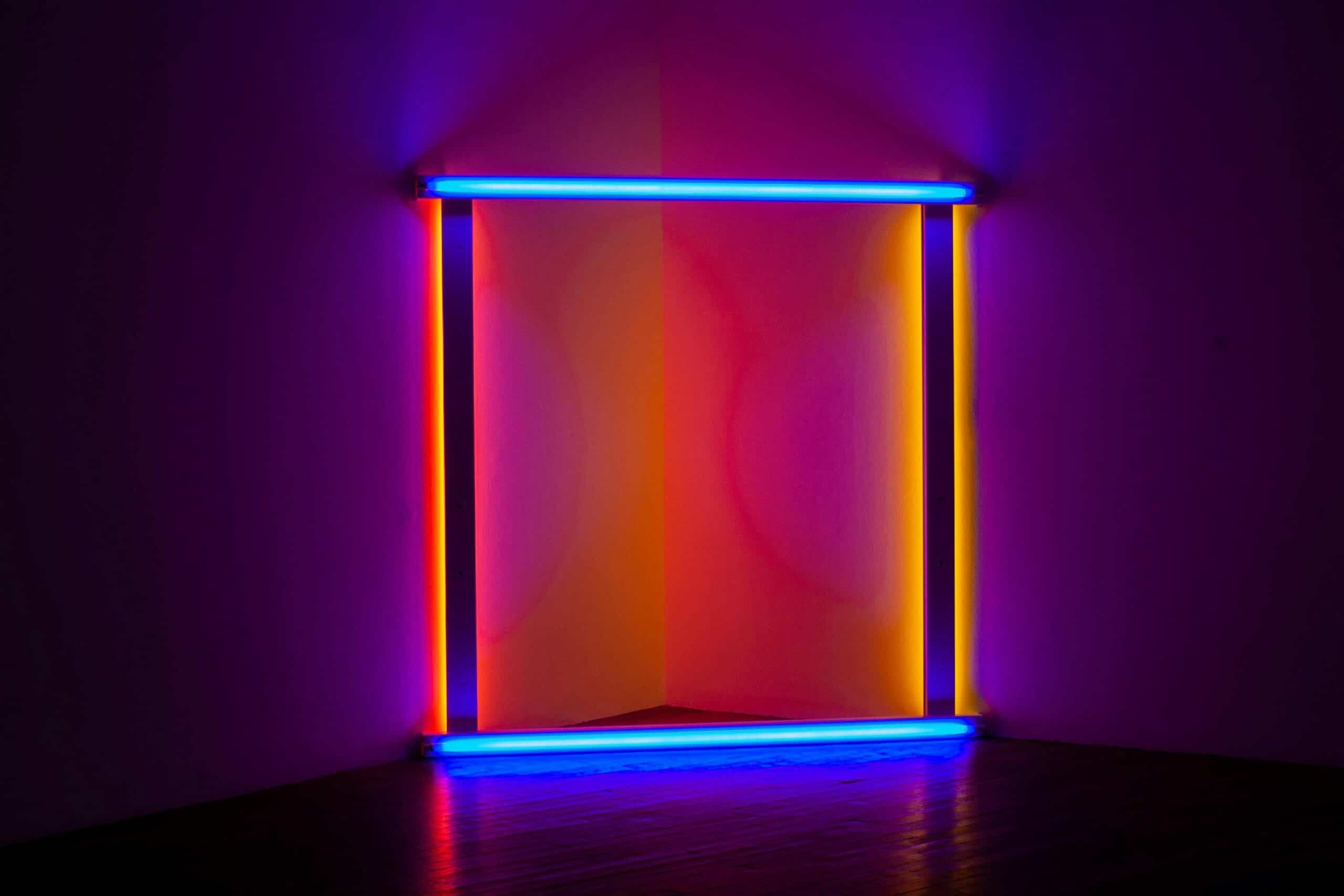 A room corner illuminated by neon lights. Blue horizontal tubes are at the top and bottom, and yellow-orange vertical tubes are on the sides, forming a glowing rectangle. The walls and dark floor reflect the vibrant colors.
