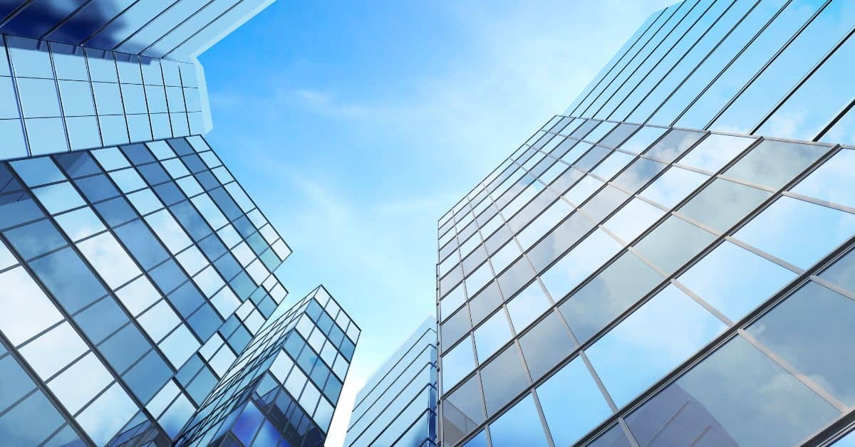 Low-angle view of multiple modern glass buildings reflecting a clear blue sky with white clouds.