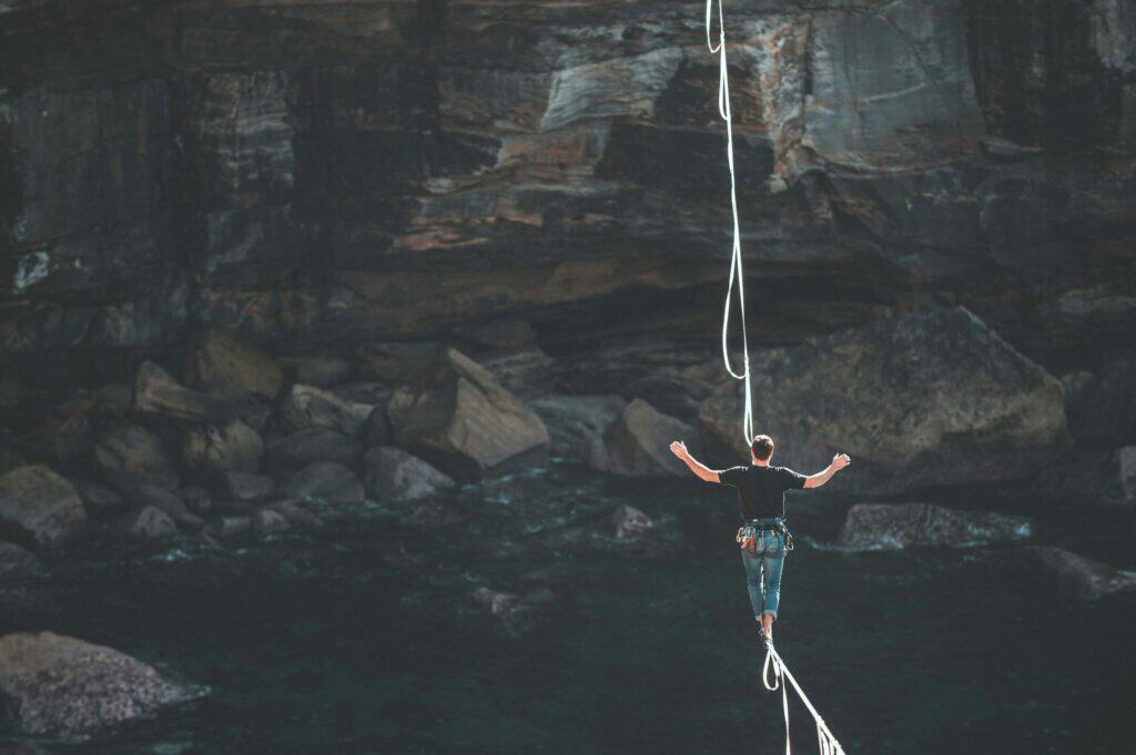 A person in a black shirt and jeans slacklining barefoot over dark water with arms outstretched for balance, against a backdrop of rocky cliffs and a cave.