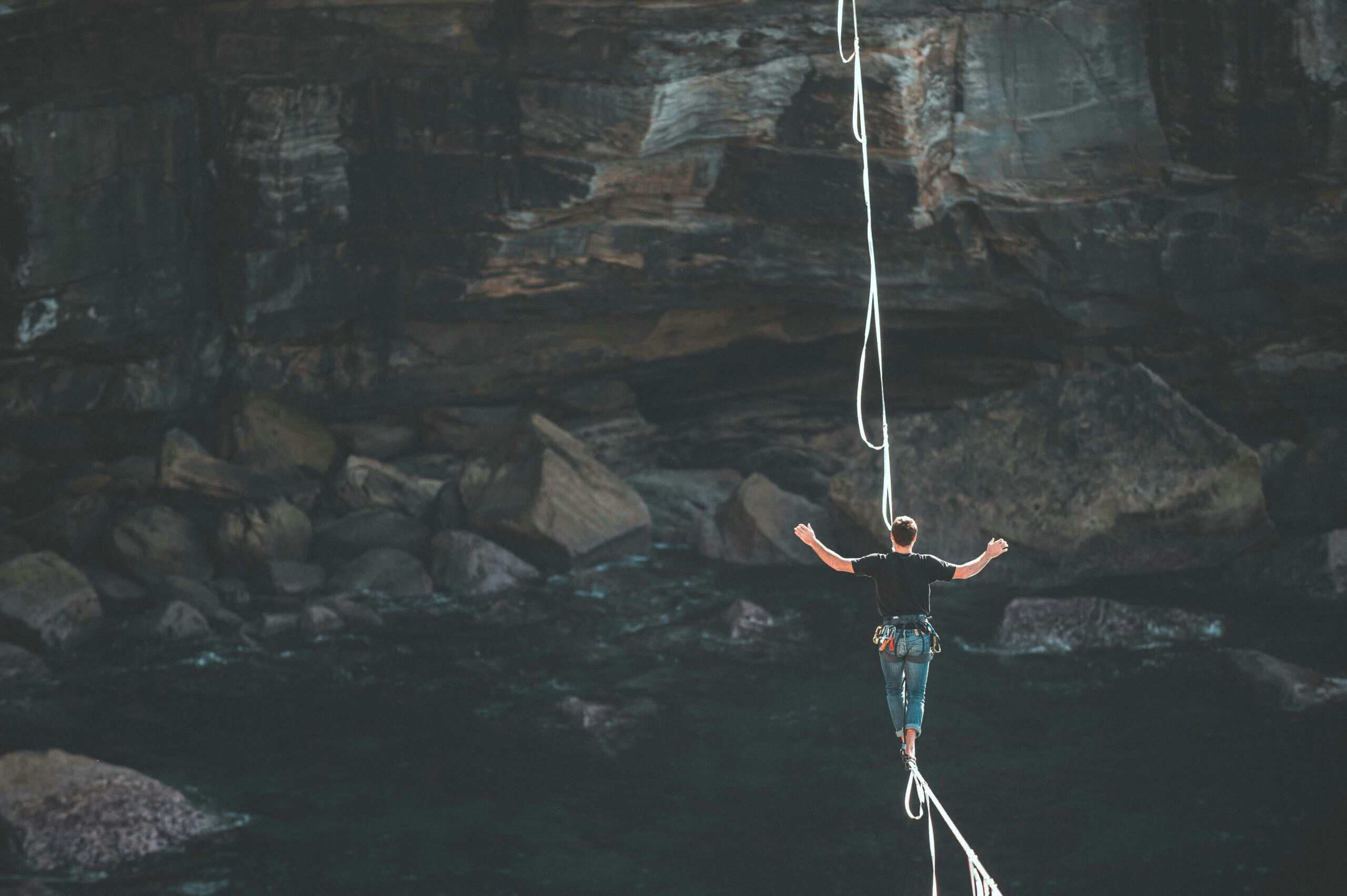A person in a black shirt and jeans slacklining barefoot over dark water with arms outstretched for balance, against a backdrop of rocky cliffs and a cave.