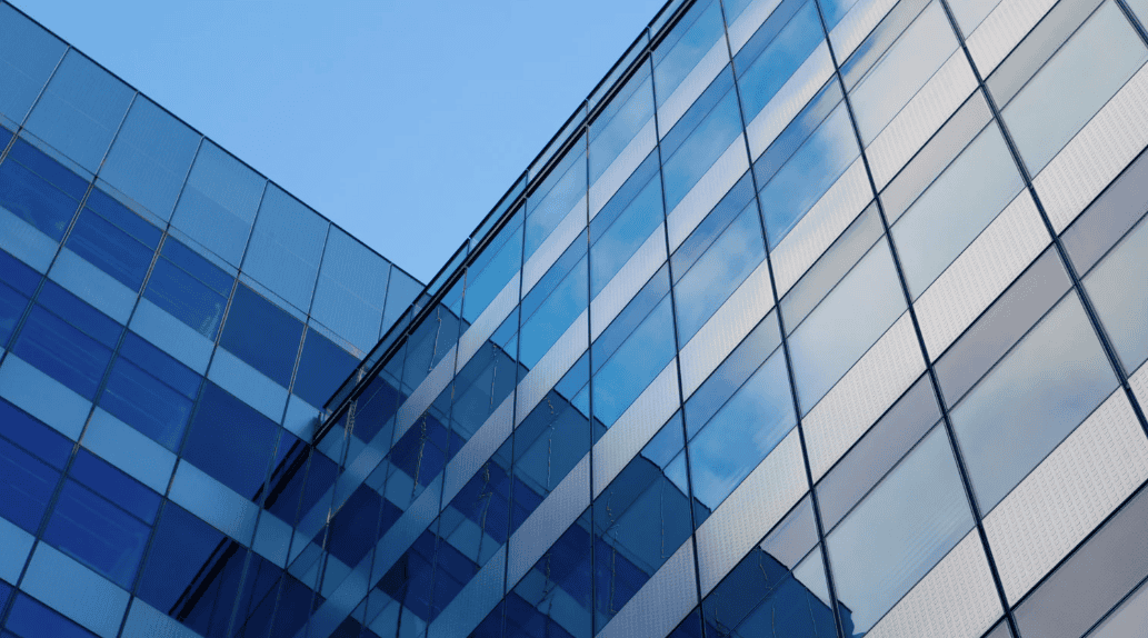 Close-up of a modern building facade with blue glass windows and light grey textured panels reflecting a clear sky.