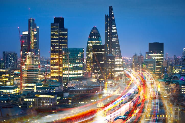 A long-exposure shot of the London skyline at night, featuring illuminated skyscrapers like The Gherkin and The Walkie-Talkie, with vibrant red and yellow light trails from traffic in the foreground.