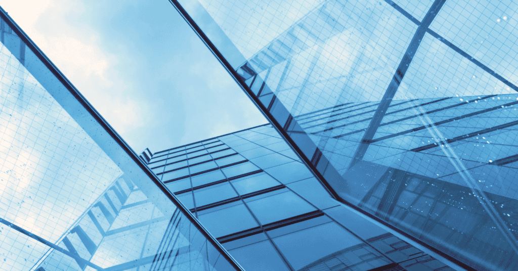 Low-angle view of a modern glass building facade reflecting a blue sky with clouds, showing geometric patterns and reflections.