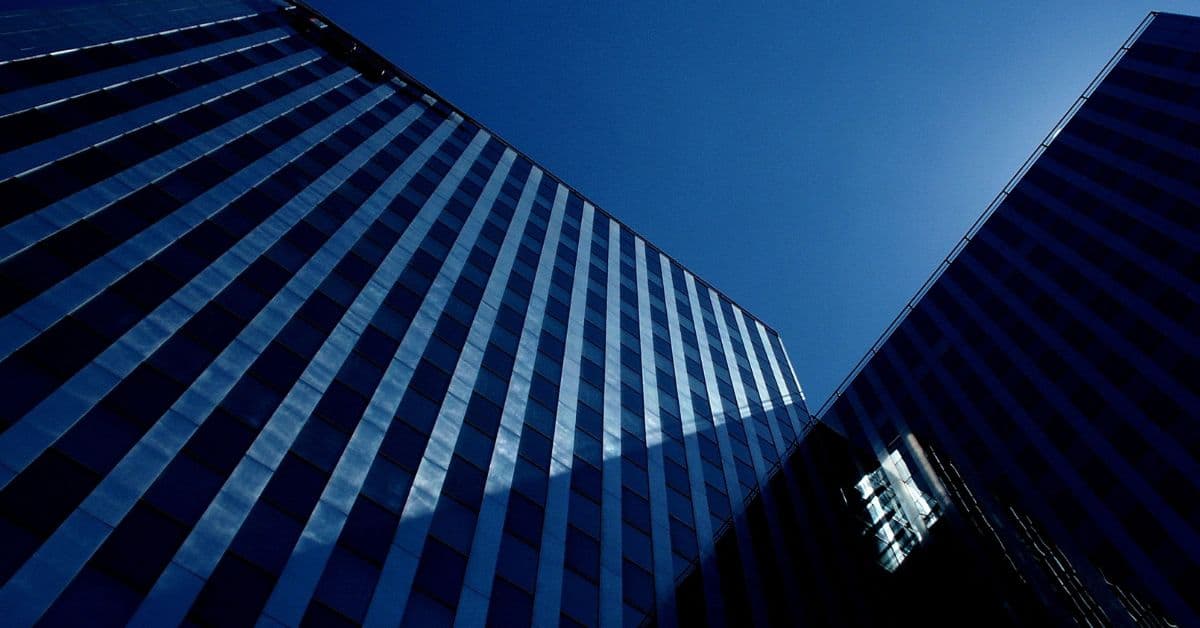 Two modern buildings with blue glass facades, viewed from below against a clear blue sky. One building is brightly lit, the other is in shadow.