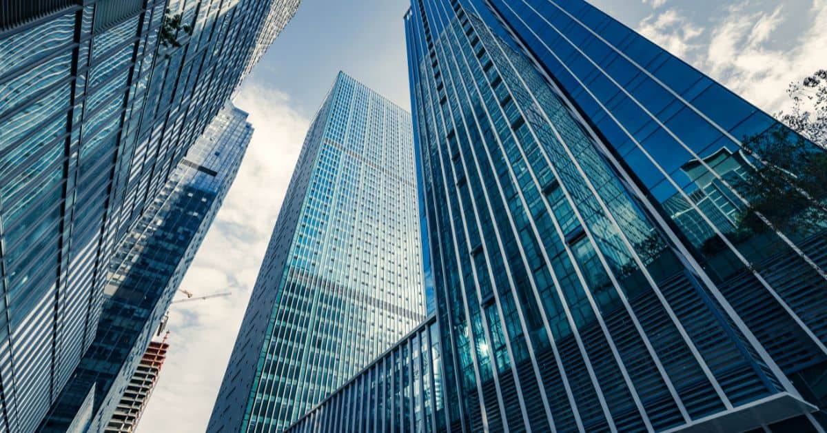 Three modern skyscrapers with blue and grey glass facades, viewed from below against a bright, cloudy sky.