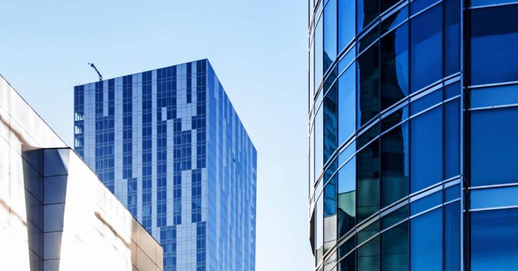 Low-angle view of three modern buildings. One on the right has a curved facade of dark blue reflective glass. Another in the center-left is rectangular with light blue and white striped panels, topped by a small crane. A third, light-colored building is partially visible on the left.