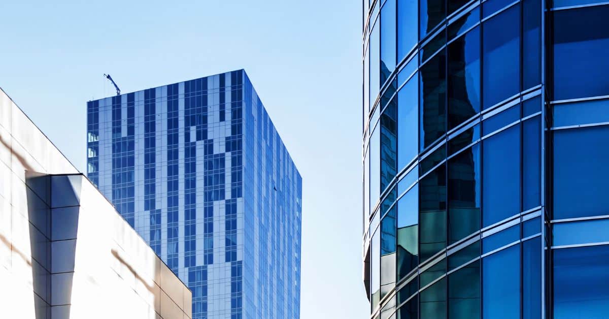 Low-angle view of three modern buildings. One on the right has a curved facade of dark blue reflective glass. Another in the center-left is rectangular with light blue and white striped panels, topped by a small crane. A third, light-colored building is partially visible on the left.