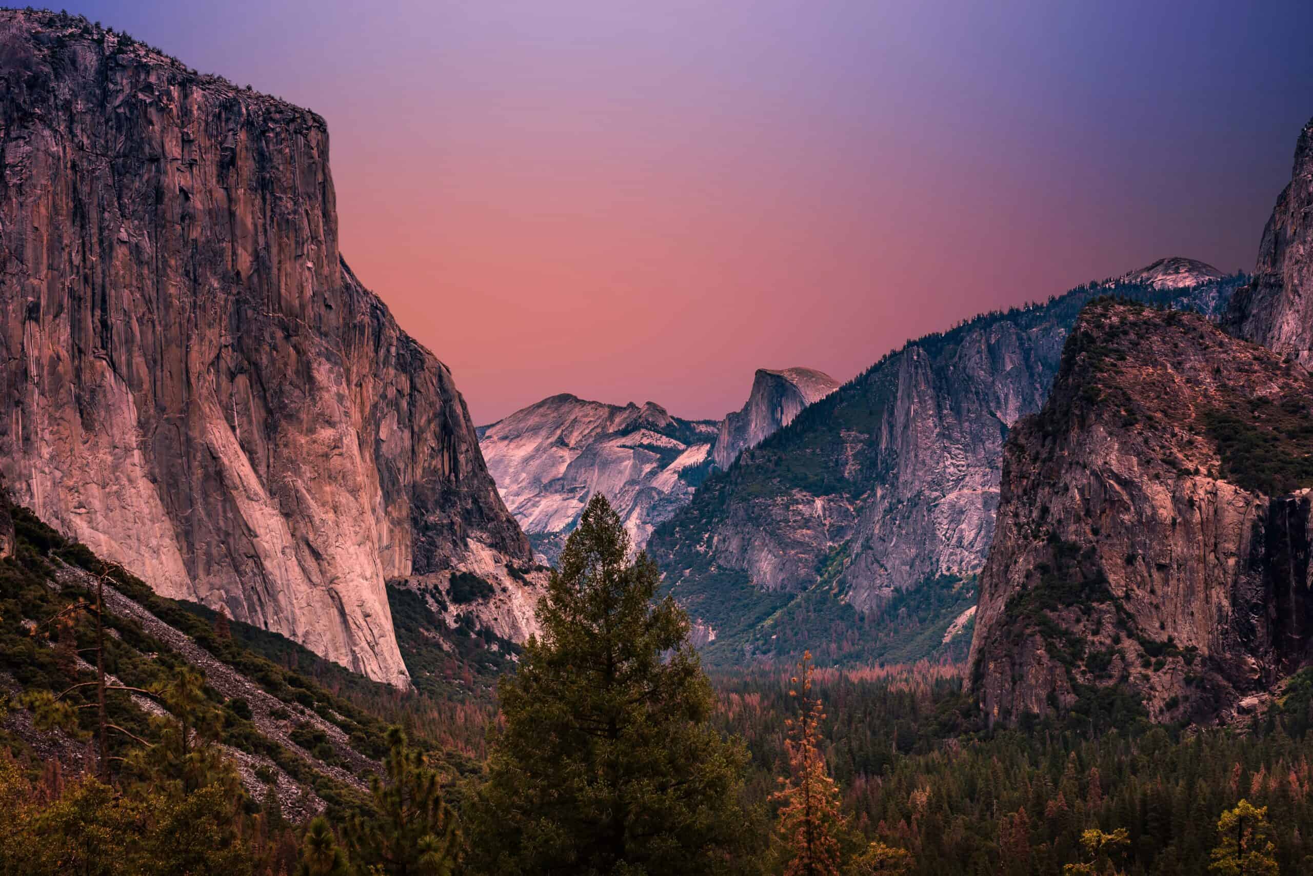 A mountain valley at twilight, with a reddish-orange to purple sky, flanked by a massive light granite cliff on the left and a dark rugged mountain on the right, with a dense forest in the valley and distant peaks.