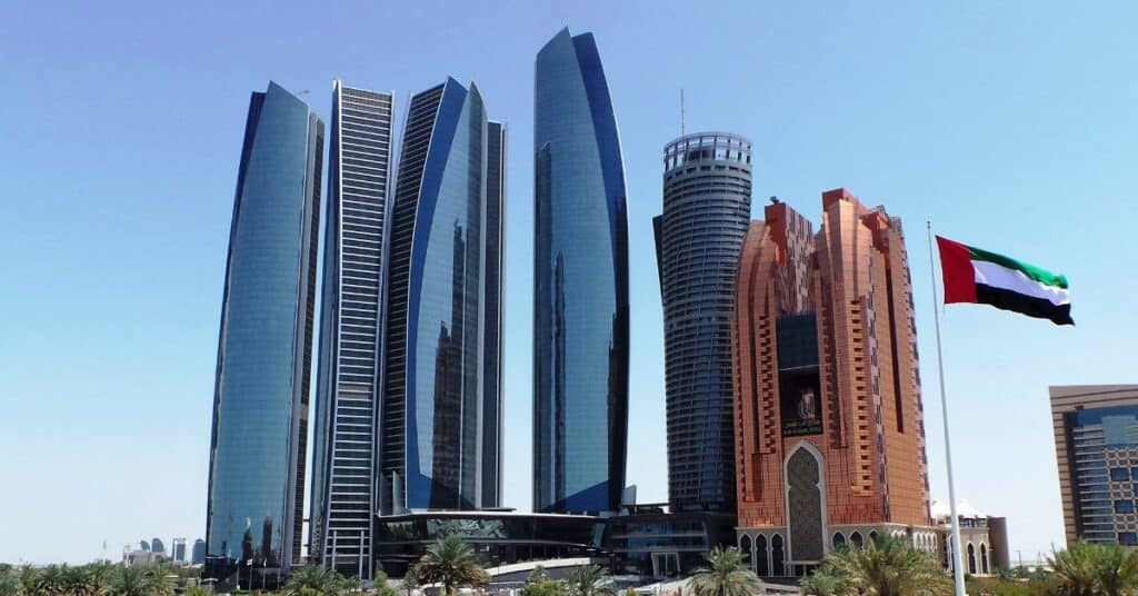 A cluster of modern skyscrapers with blue glass and reddish-brown facades, with the UAE flag flying on a flagpole in the foreground, under a clear blue sky.