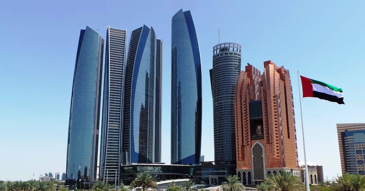A cluster of modern skyscrapers with blue glass and reddish-brown facades, with the UAE flag flying on a flagpole in the foreground, under a clear blue sky.