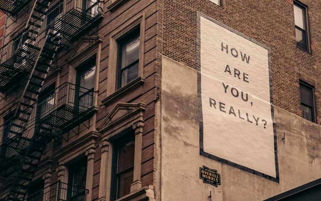 A mural on a brick building reads "HOW ARE YOU, REALLY?" with a fire escape visible on an adjacent wall.