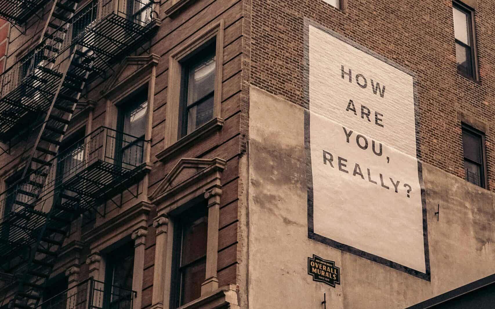 A mural on a brick building reads "HOW ARE YOU, REALLY?" with a fire escape visible on an adjacent wall.