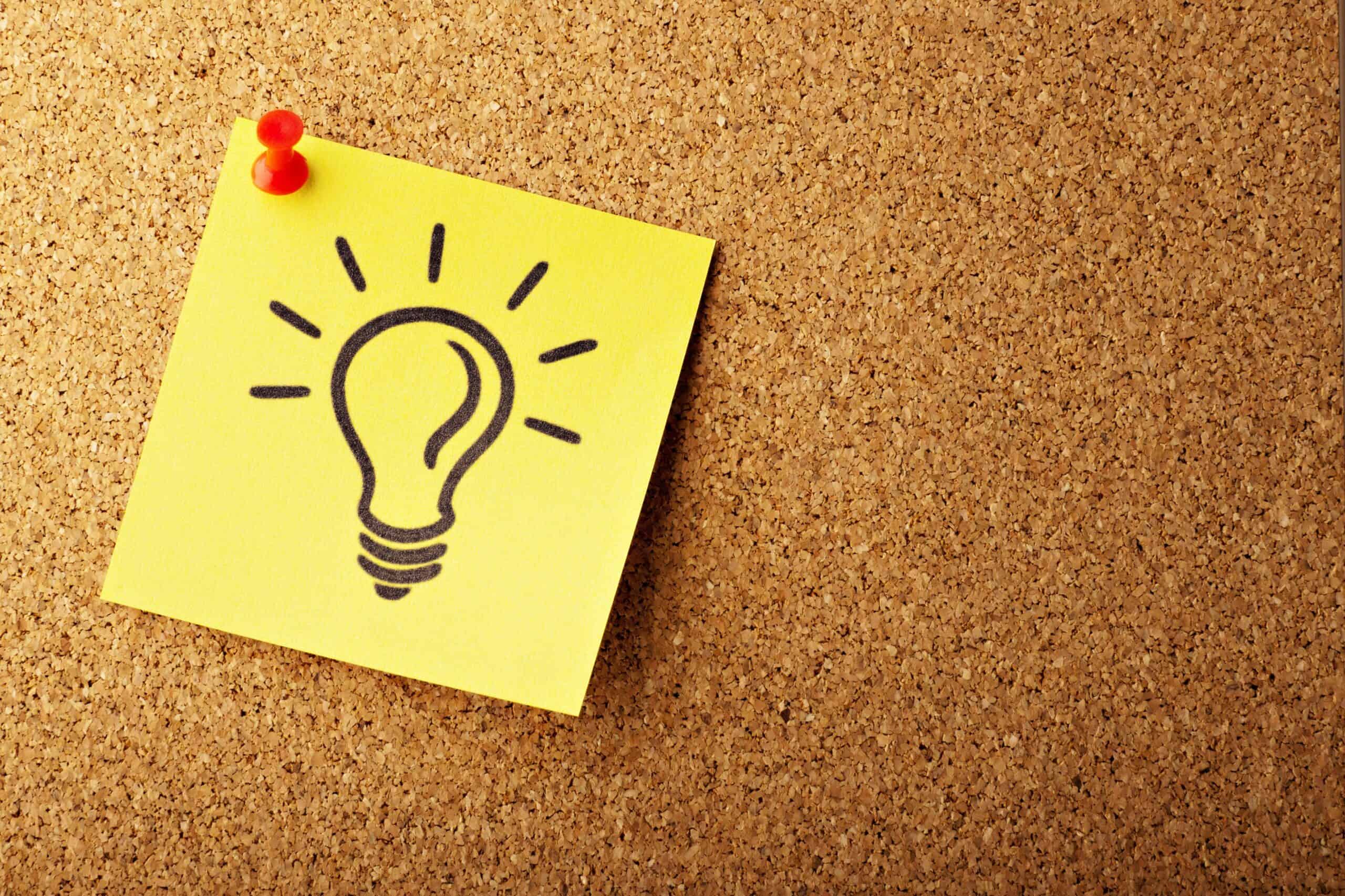 A yellow sticky note with a black lightbulb drawing pinned to a cork board with a red pushpin.
