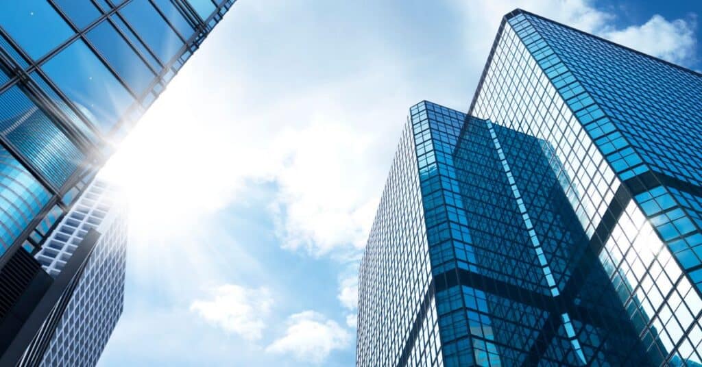 Low-angle view of three modern skyscrapers with blue glass facades, reflecting the bright sun and a blue sky with white clouds.