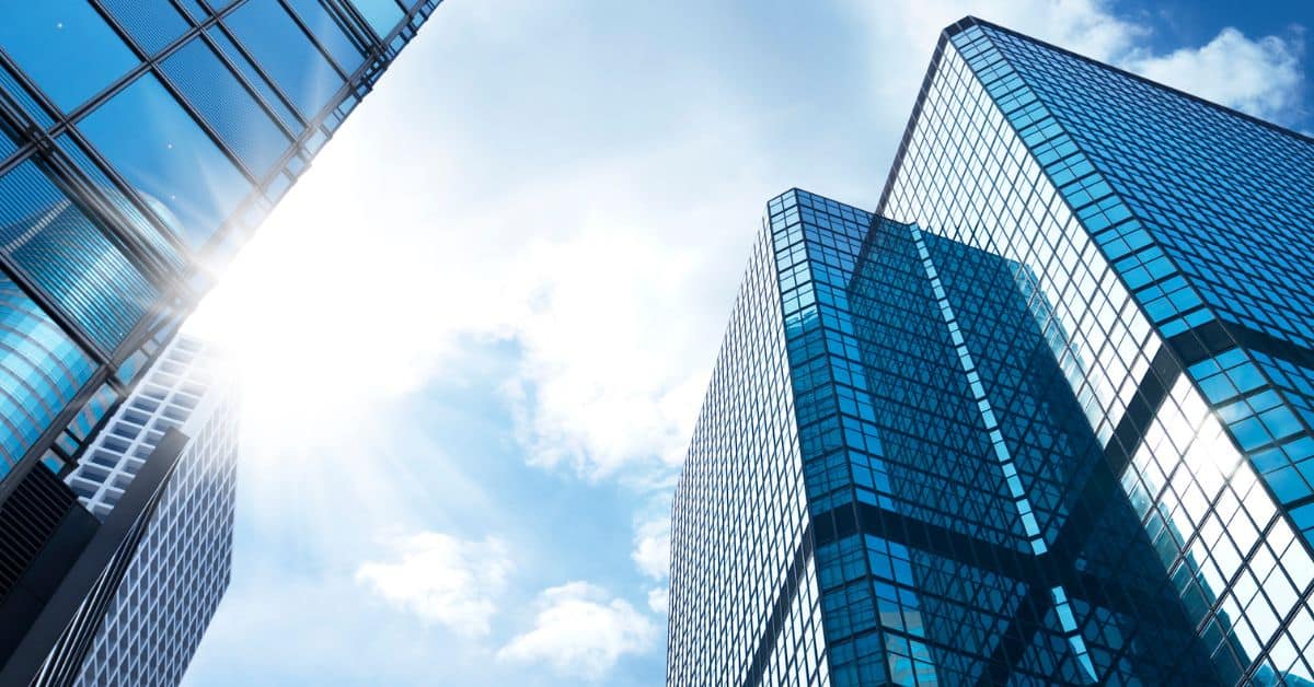 Low-angle view of three modern skyscrapers with blue glass facades, reflecting the bright sun and a blue sky with white clouds.