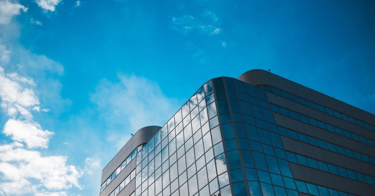 Upward view of a modern building with a curved, glass facade reflecting a bright blue sky with white clouds.