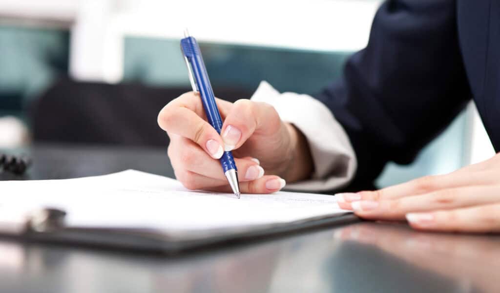 Close-up of a person's hands, with manicured nails, writing on a white document with a blue pen.