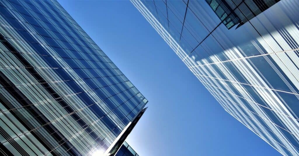 Upward view of two modern glass buildings against a clear blue sky, showing reflective facades and geometric window patterns.