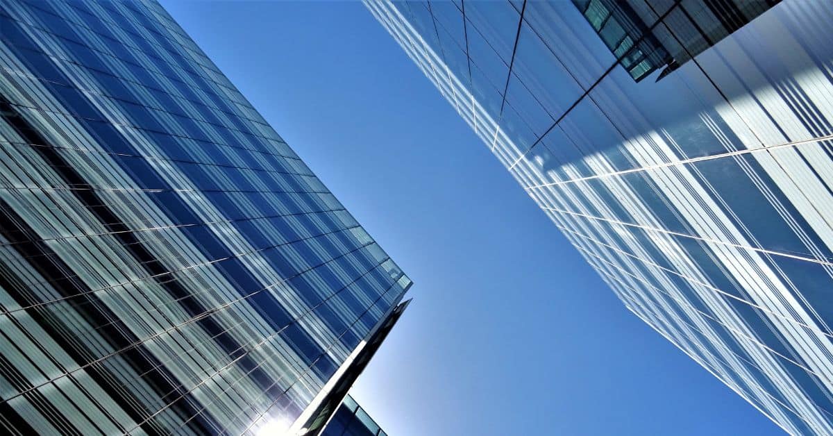 Upward view of two modern glass buildings against a clear blue sky, showing reflective facades and geometric window patterns.