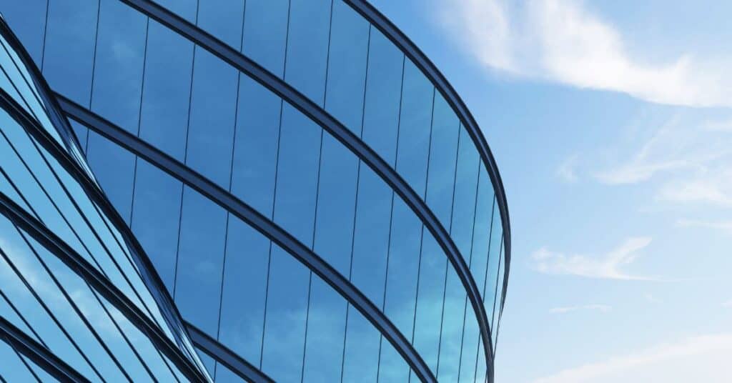 Close-up of a modern, curved building facade made of blue reflective glass, set against a bright blue sky with white clouds.