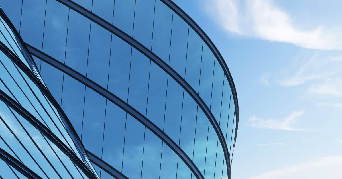Close-up of a modern, curved building facade made of blue reflective glass, set against a bright blue sky with white clouds.