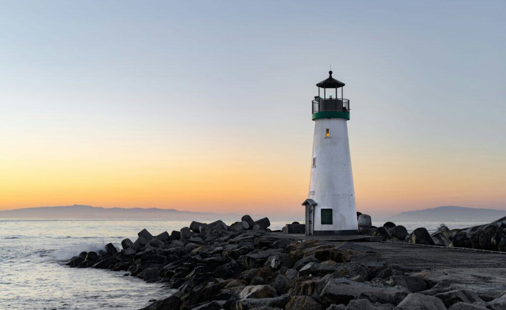 A white lighthouse with a green top on a rocky breakwater at sunrise or sunset, with a gradient sky and distant mountains.