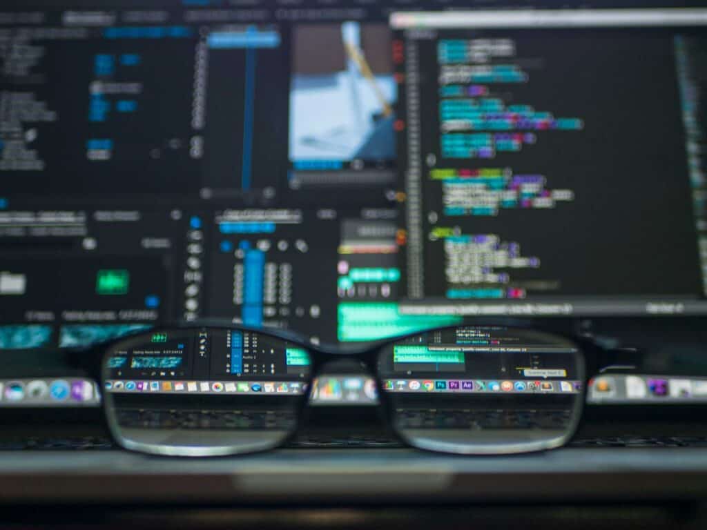 Black-framed eyeglasses rest on a laptop keyboard, reflecting and refracting a blurred screen displaying code and video editing software.