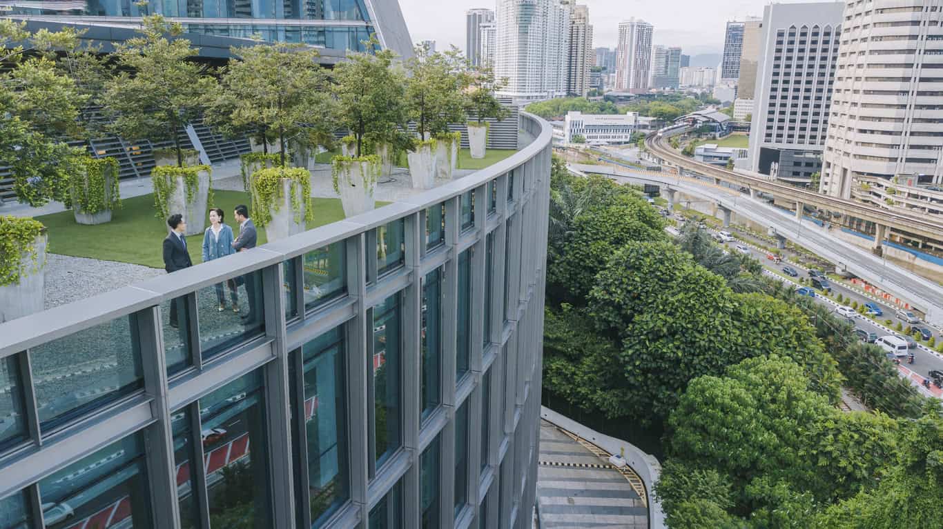 Three business people on a green rooftop garden with trees, overlooking a city with tall buildings, roads, and train tracks.