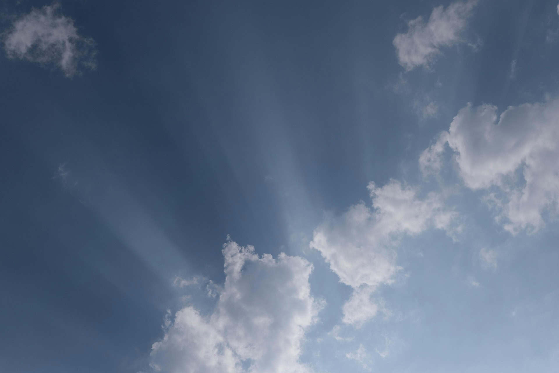 A blue sky with white and grey clouds, from which crepuscular rays of sunlight fan out.