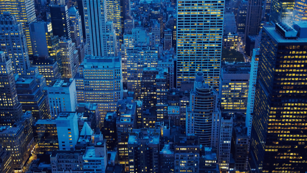 Aerial view of a dense city at night, with numerous skyscrapers and buildings featuring many illuminated windows.