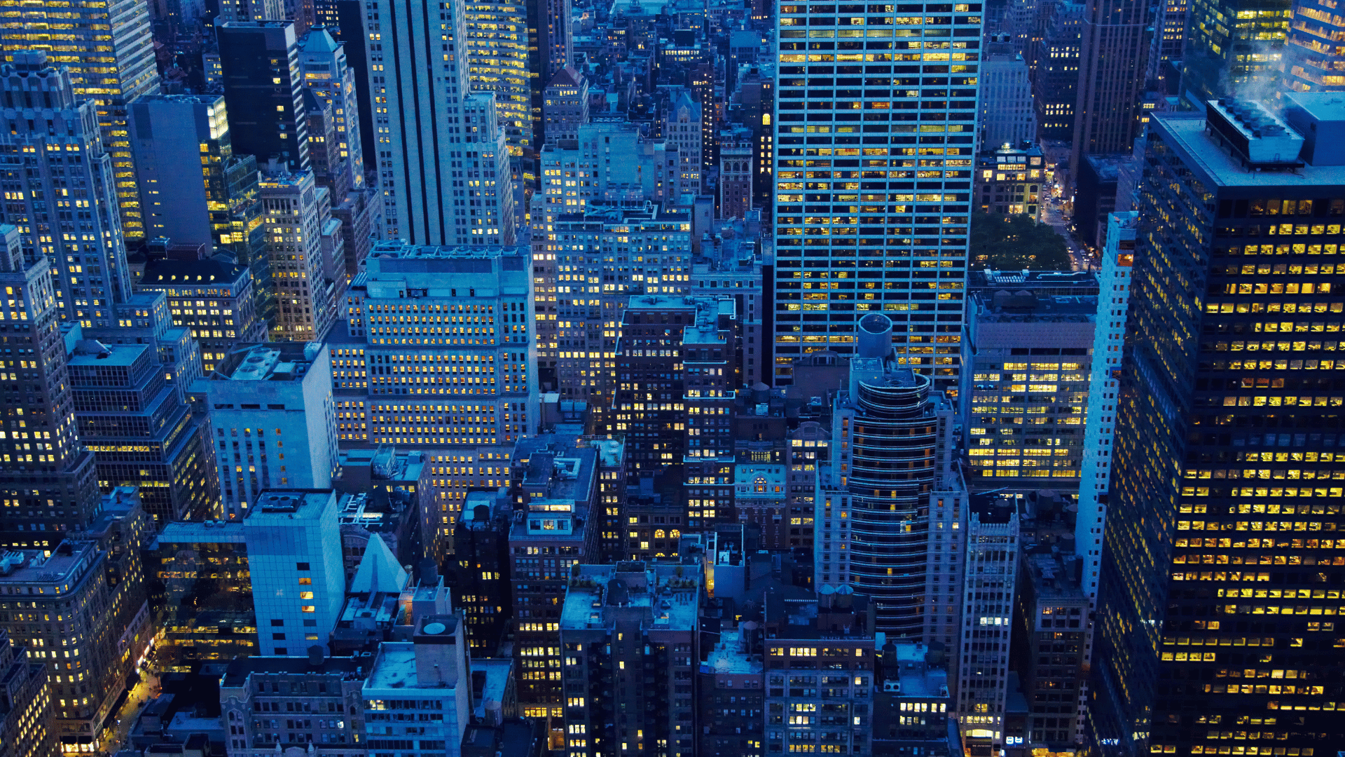 Aerial view of a dense city at night, with numerous skyscrapers and buildings featuring many illuminated windows.
