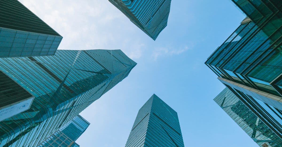 Low-angle view of multiple modern skyscrapers with blue-green glass facades against a clear blue sky.