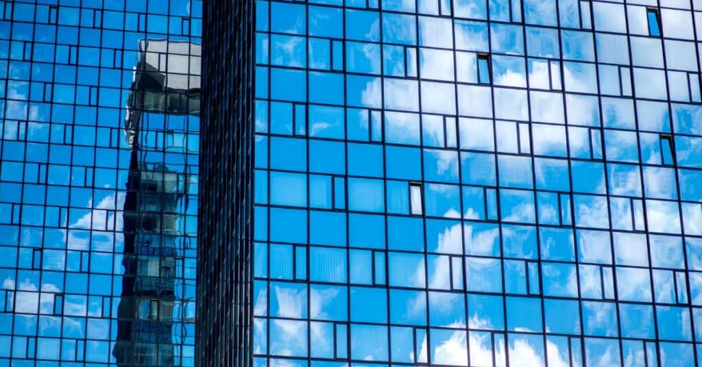 A modern glass building facade reflecting a blue sky with white clouds and a distorted image of another building.