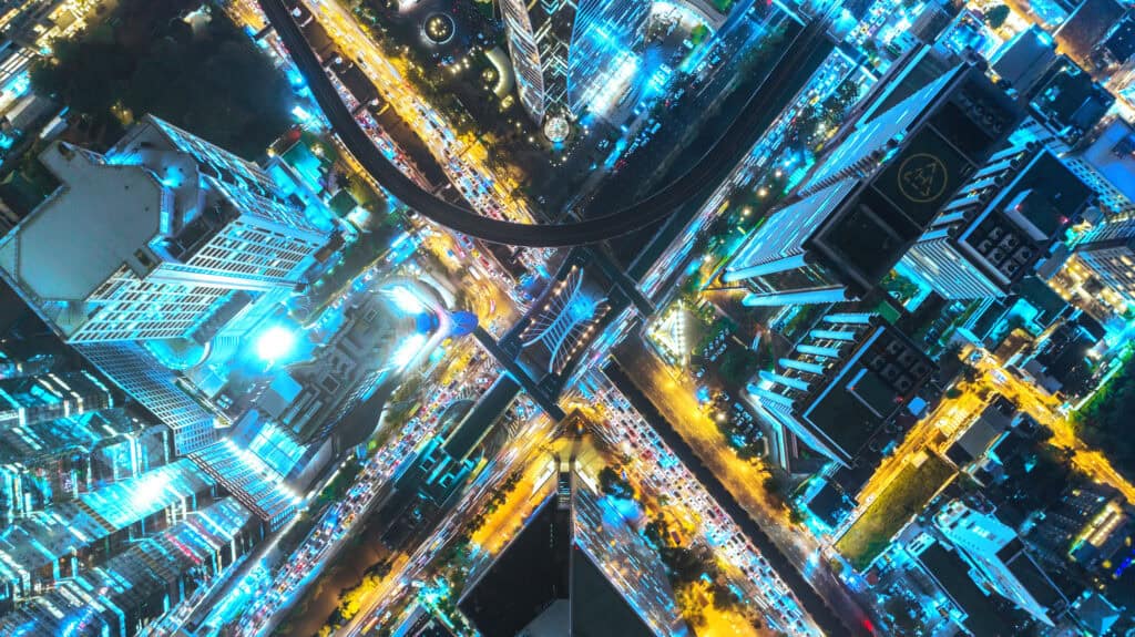 Aerial top-down view of a city at night, showing high-rise buildings illuminated with blue lights and roads filled with streaking yellow and red traffic lights.