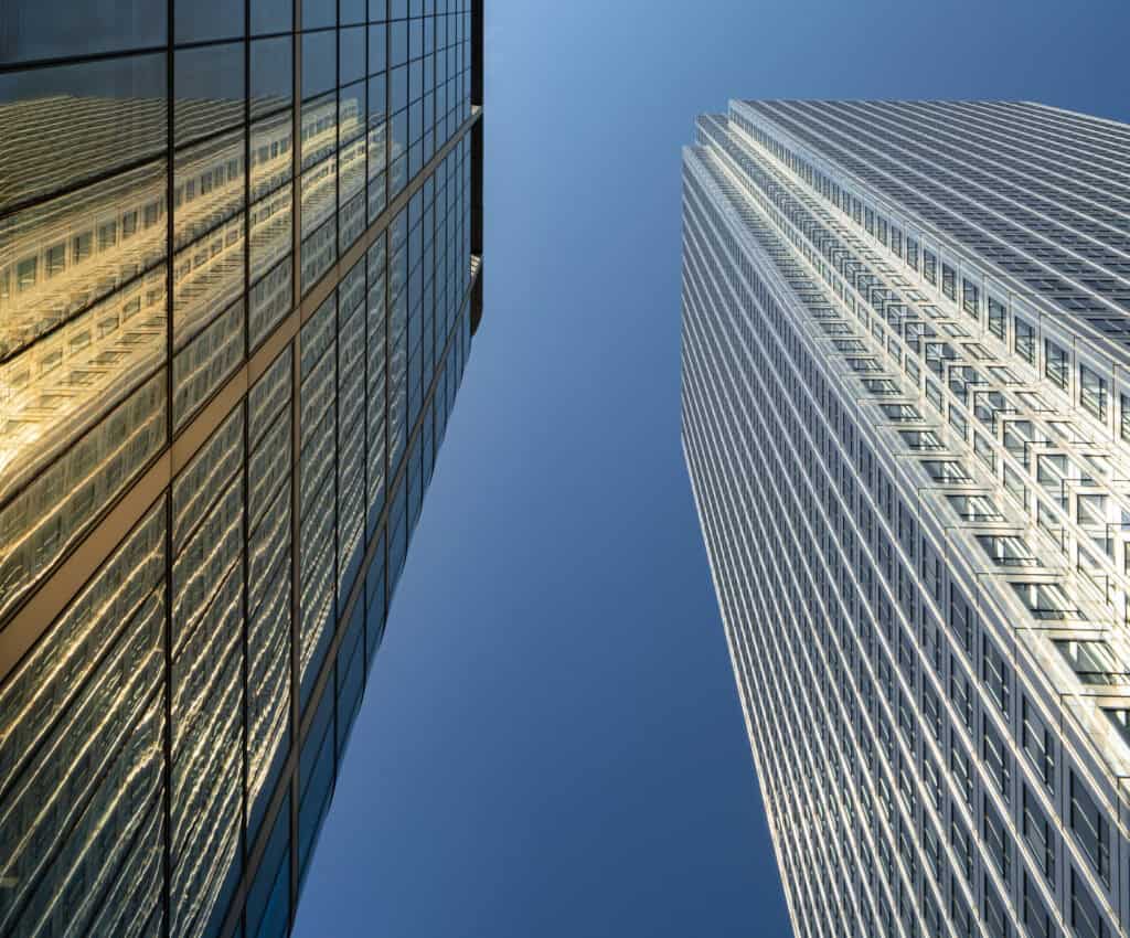 Two tall skyscrapers against a clear blue sky, one dark glass reflecting golden light, the other with a light grid pattern.