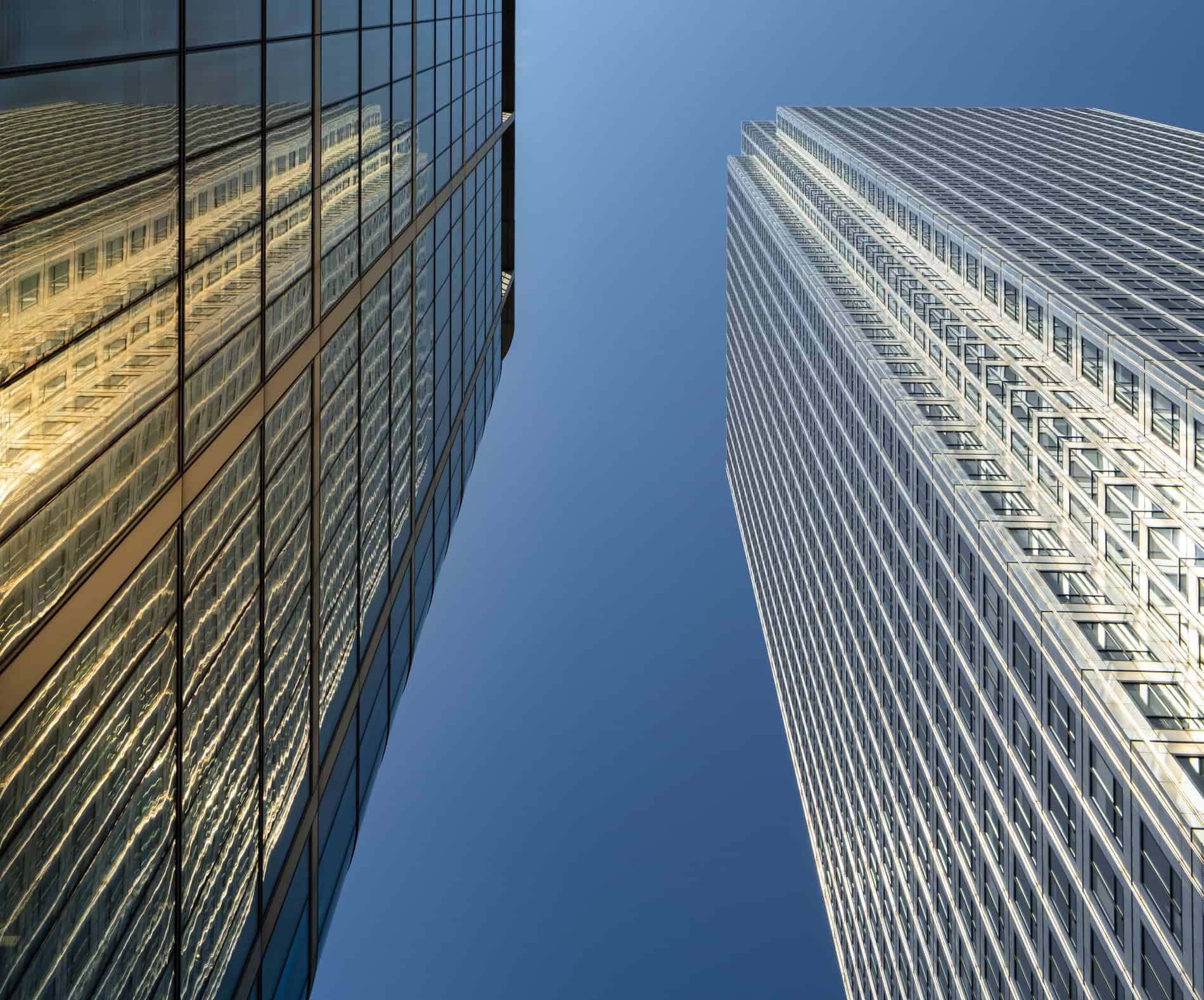 Two tall skyscrapers against a clear blue sky, one dark glass reflecting golden light, the other with a light grid pattern.