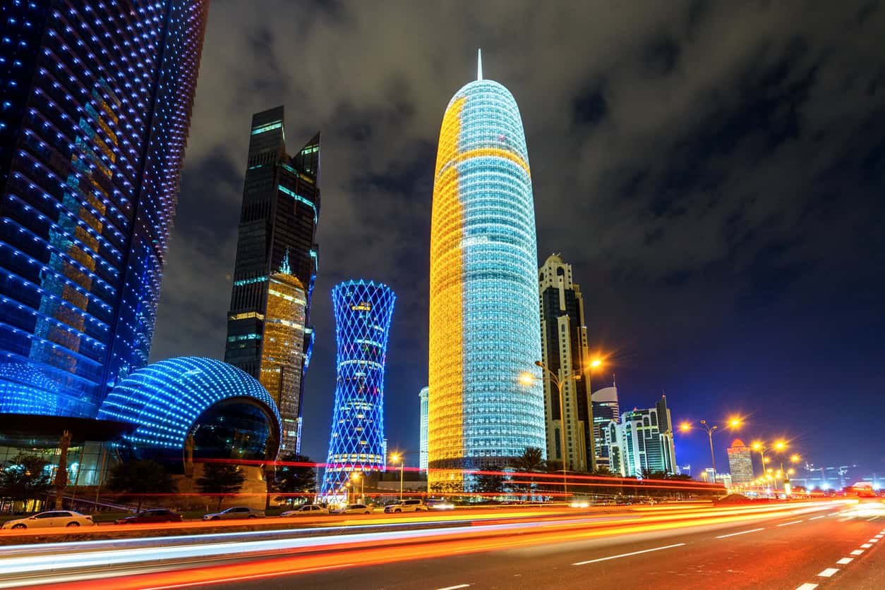 Night view of a modern city skyline with brightly lit skyscrapers and long exposure light trails from cars on a multi-lane road.