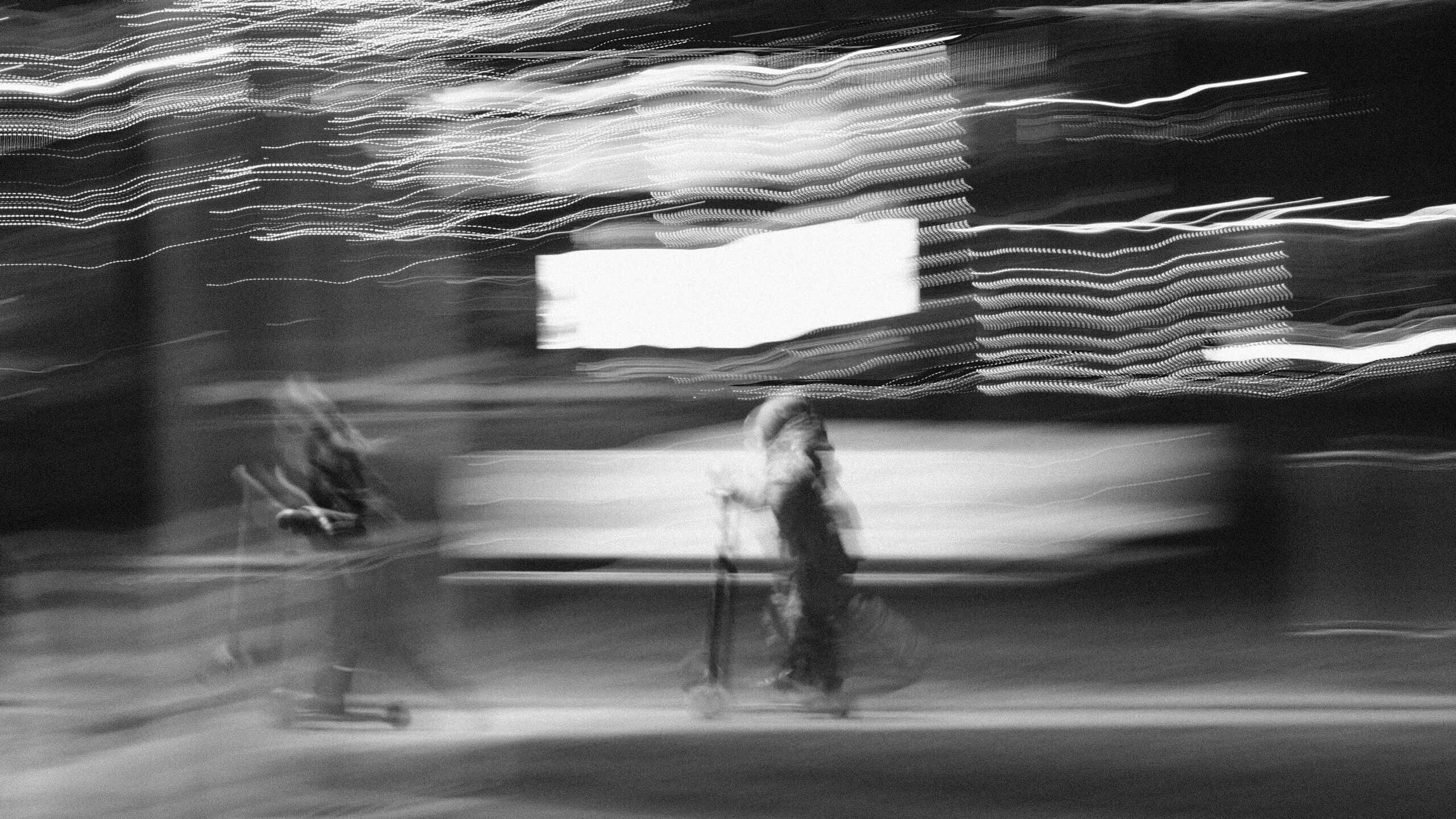 Black and white image with heavy motion blur showing two indistinct figures on scooters moving past streaks of light.