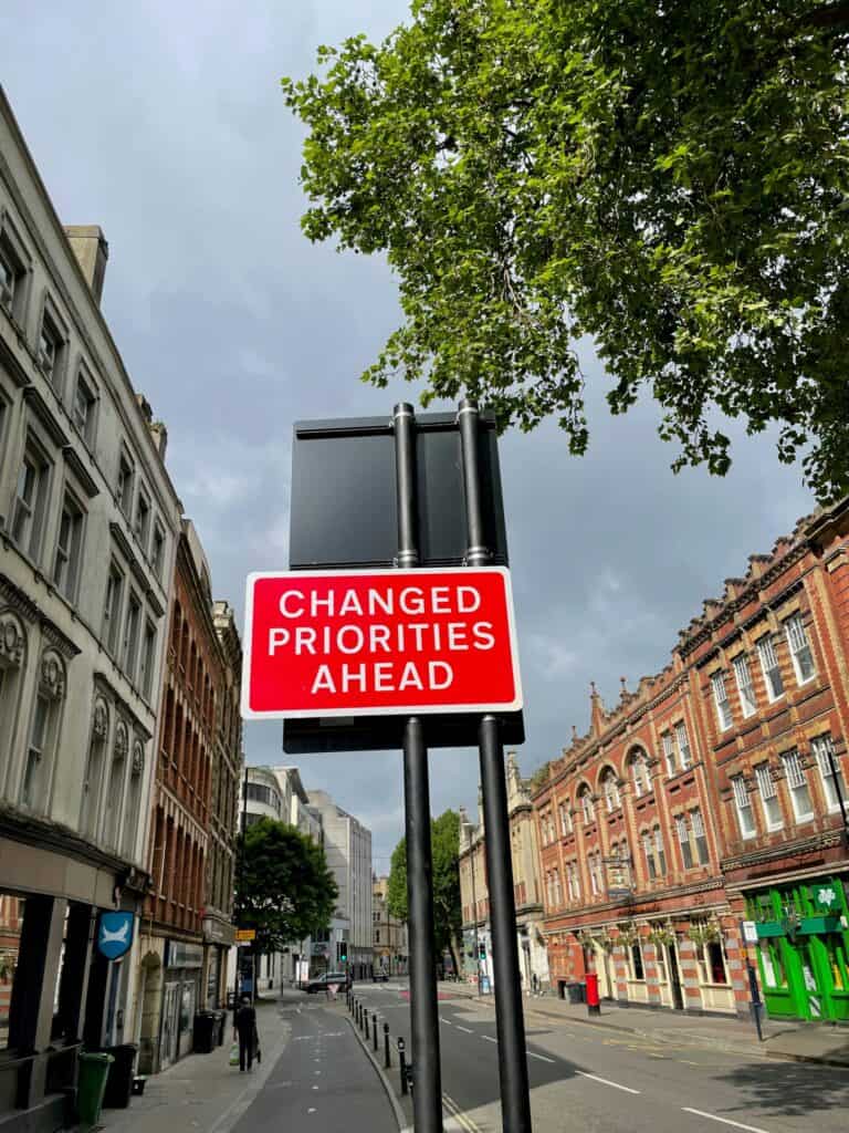 A red road sign with white text "CHANGED PRIORITIES AHEAD" on a city street with brick and stone buildings, trees, and an overcast sky.