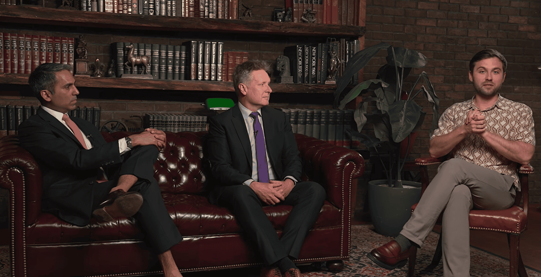 Three men in a room with a bookshelf. One in a suit and pink tie, one in a suit and purple tie, and one in a patterned shirt.