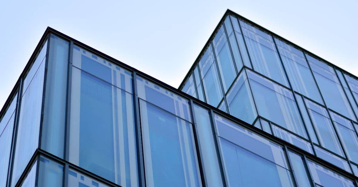 Upward view of a modern building with blue glass facade and dark frames against a light blue sky.