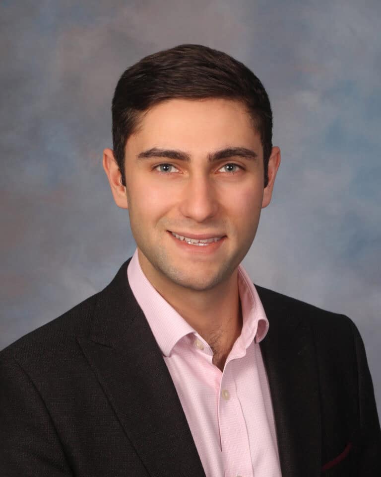 Headshot of a smiling man with dark hair and light eyes, wearing a pink shirt and dark blazer against a blue-grey background.