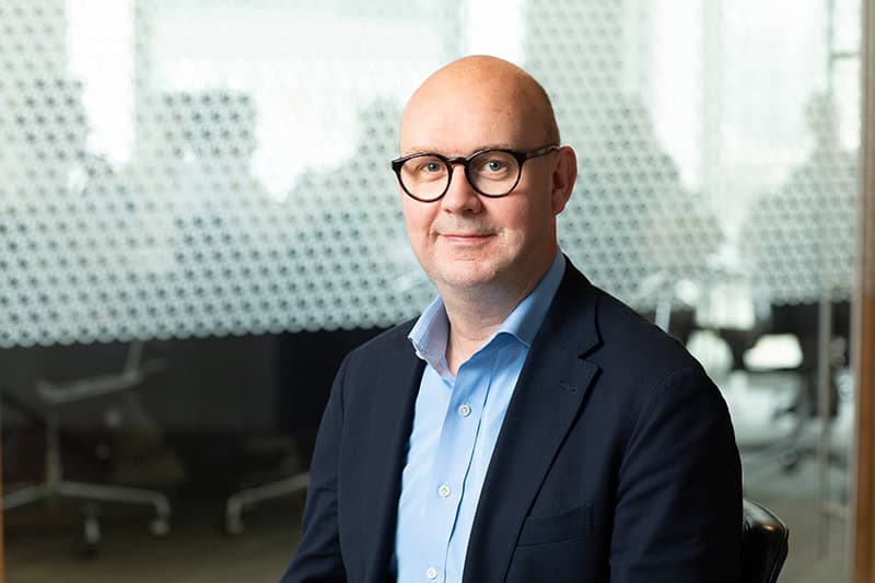 A bald man with glasses, wearing a blue shirt and dark suit jacket, smiles at the camera in an office setting.