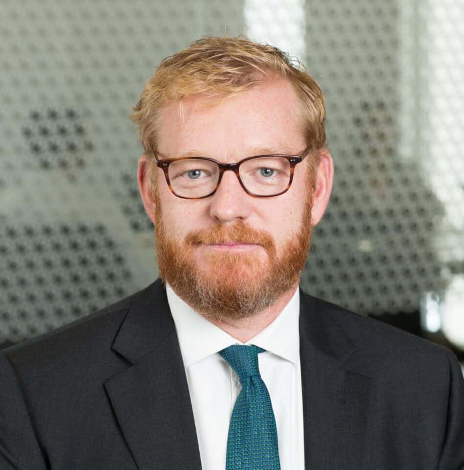 Headshot of a man with reddish-blonde hair and beard, wearing glasses, a dark suit, and a teal tie.