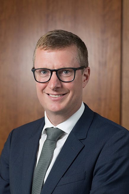 Headshot of a man with short brown hair, glasses, and a gray tie, smiling.