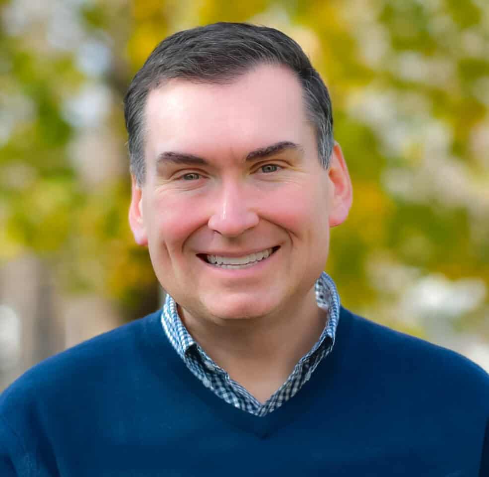 Headshot of a smiling man in a blue sweater and checkered shirt, with a blurred yellow and green leafy background.