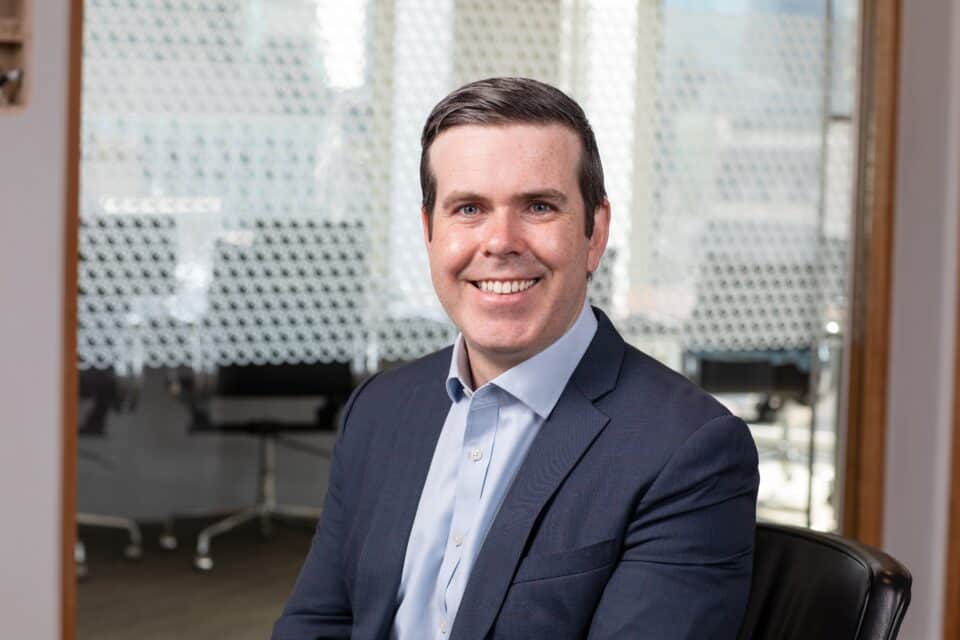 Headshot of a smiling man in a dark suit and light blue shirt, seated in an office with a patterned glass partition in the background.